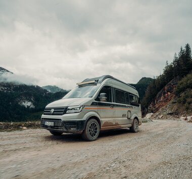Carado CV595 4x4 X-EDITION fährt über eine Schotterpiste vor einem Bergmassiv