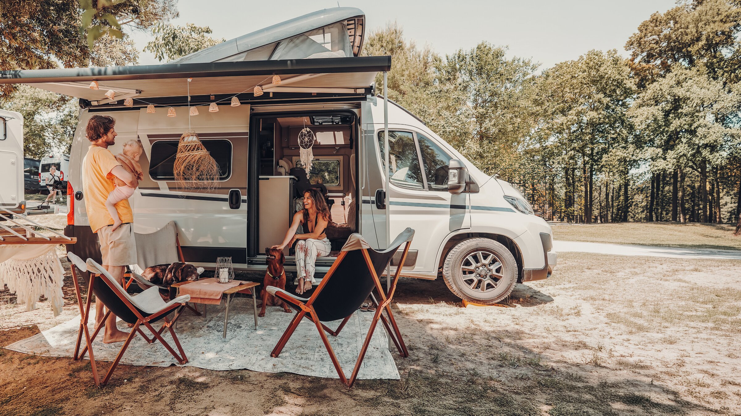 Familie mit Hund vor Carado Wohnmobil auf Campingplatz, Markise und Aufstelldach geöffnet