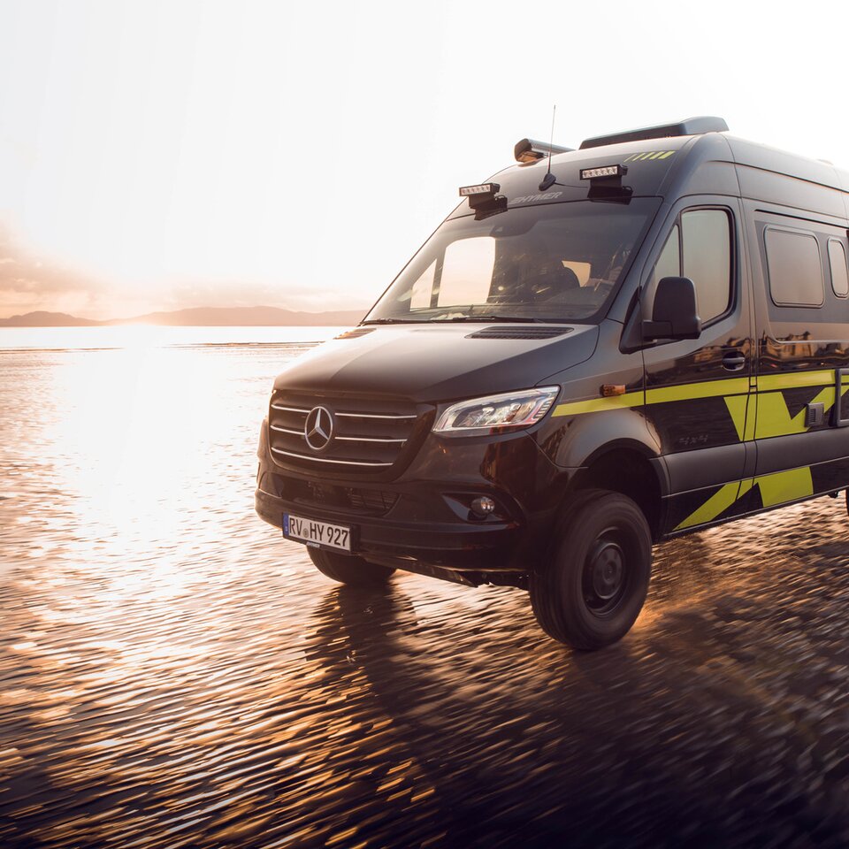 Mercedes firehjulstræk-autocamperen HYMER Grand Canyon S kører hurtigt på en solrig, våd strand. Omgivelserne er slørede på grund af dynamik.