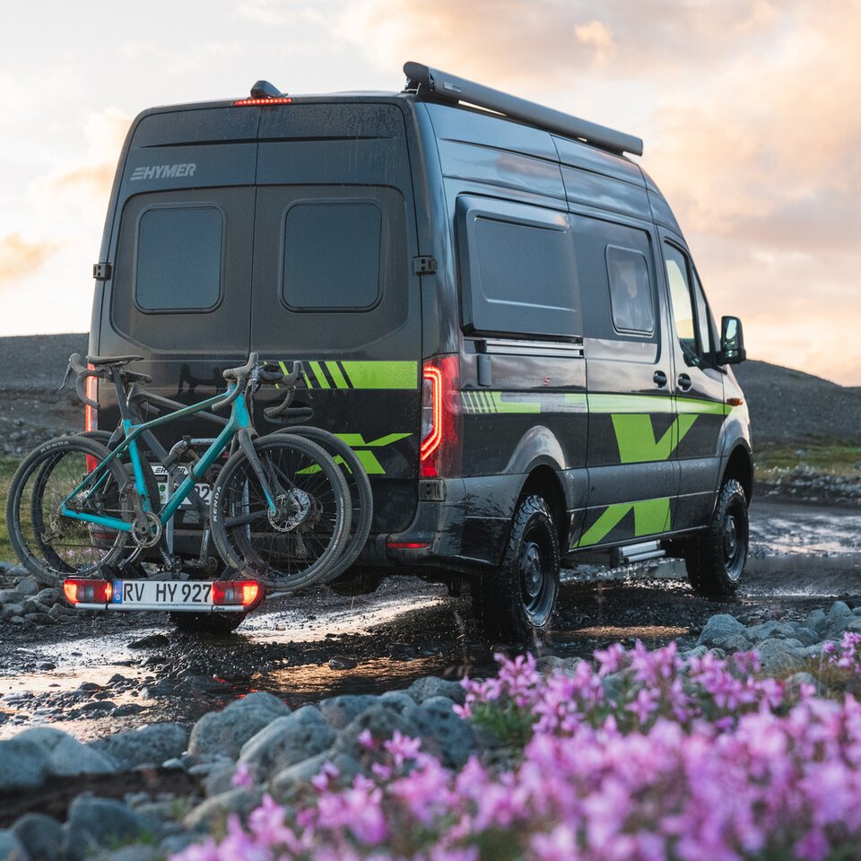 Sprinter-4x4 autocamperen HYMER Grand Canyon S CrossOver står på en grusvej med blomster og græs, cykler står på cykelholderen.