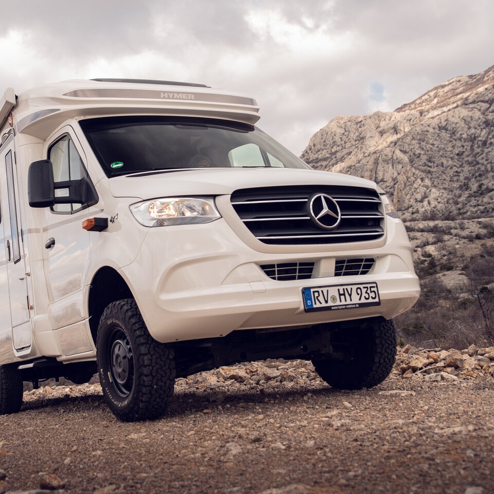 Den hvide Mercedes firehjulstræk-autocamper HYMER ML-T står på en stigende stenvej med stenede bakker og let overskyet himmel i baggrunden.