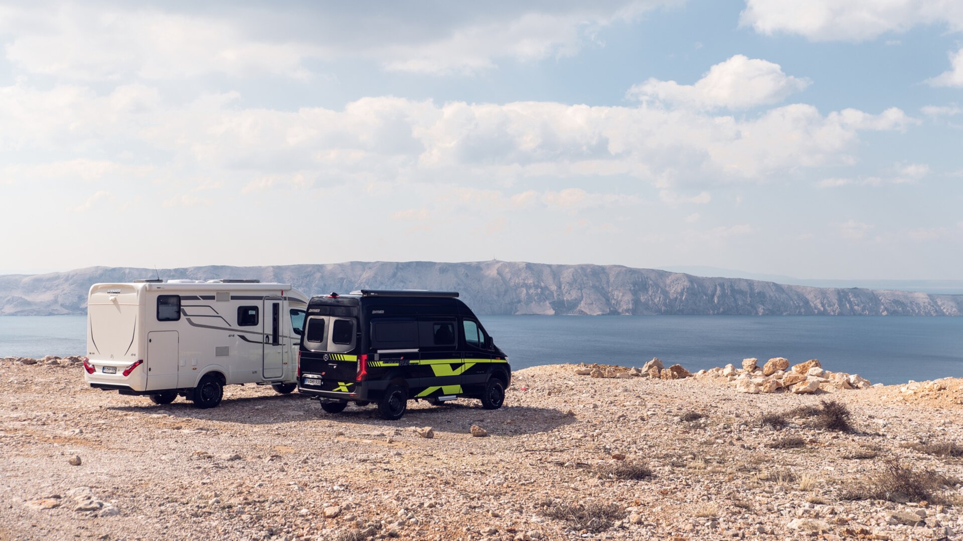 To terræn-campingbiler i hvid samt sort-grøn står på en solbeskinnet slette over havet, klipper er synlige i baggrunden.