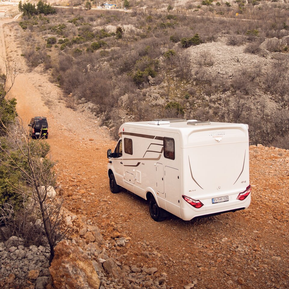To 4x4 terræn-campingbiler fra HYMER med Mercedes-førerhus kører på en bakket vej gennem ujævnt, stenet terræn med buske.