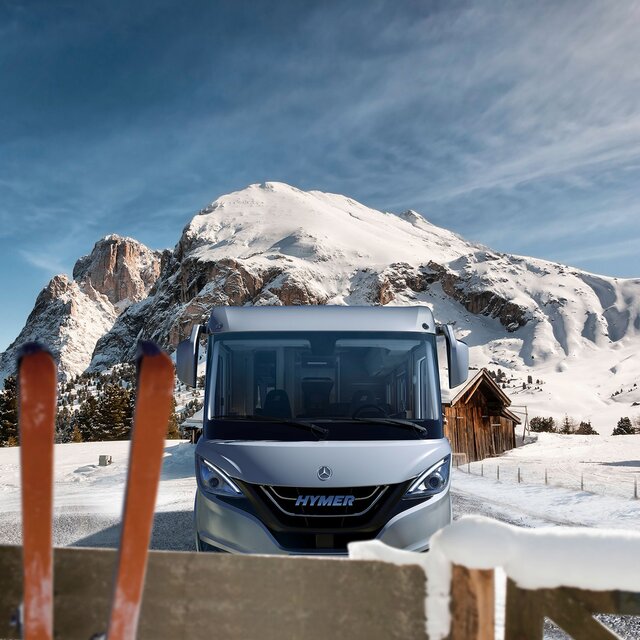 HYMER B-Klasse MasterLine in zonnig, alpine winterlandschap met houten schuur en een paar ski's tegen een houten hek