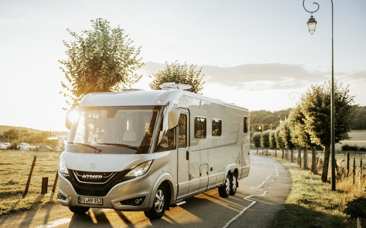 HYMER B-Klasse MasterLine I dubbelassig voertuig rijdt op een met bomen omgeven weg in een warme lichte atmosfeer