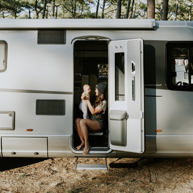 Jonge vrouw met baby zittend bij de ingang van een HYMER-camper met de motorklep open en de vuilnisbak geïntegreerd.