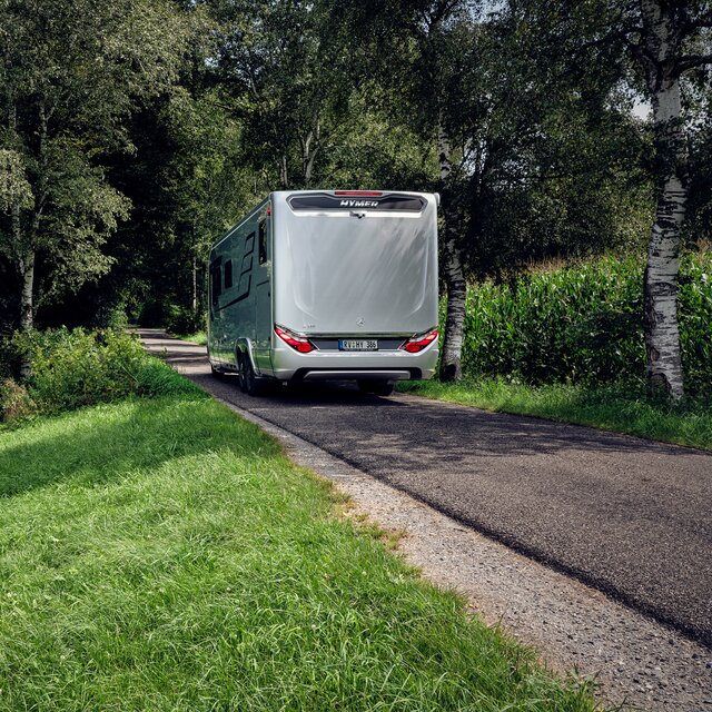 HYMER camper B-ML I 880 van achteren op de weg aan de rand van het bos met groene weide en maïsveld