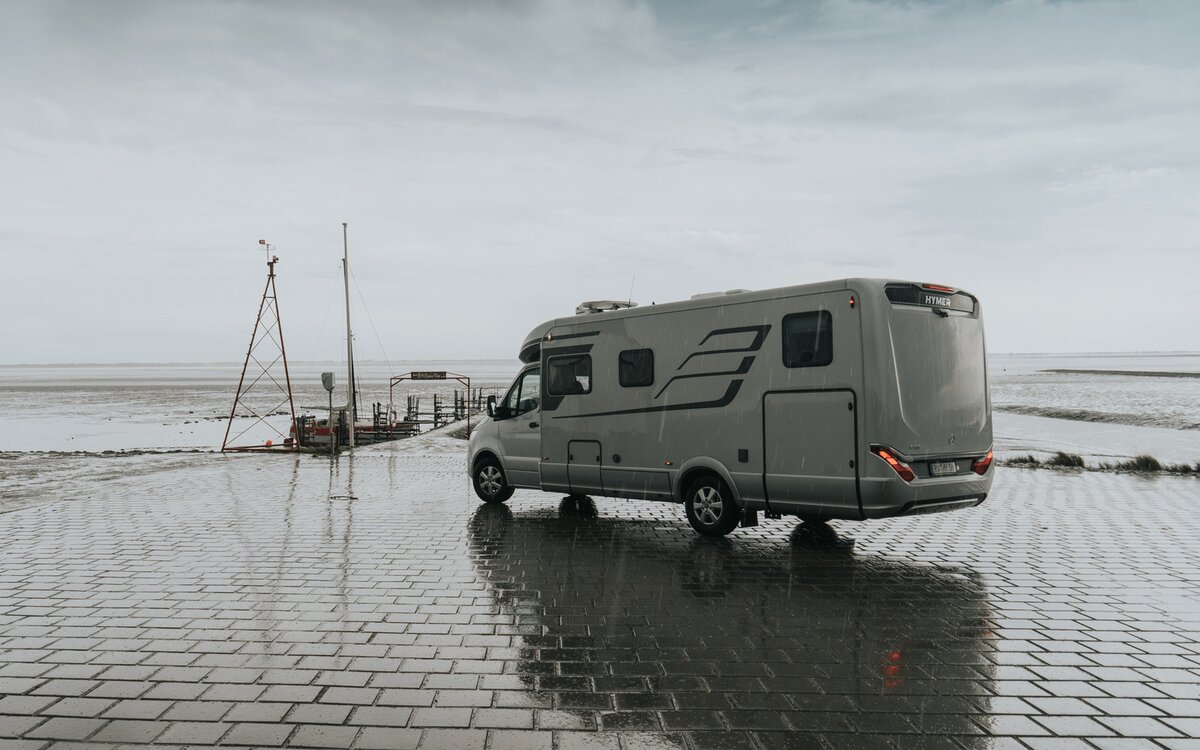 Partially integrated HYMER motorhome (rear/side view) stands on a paved surface by the sea in rainy weather