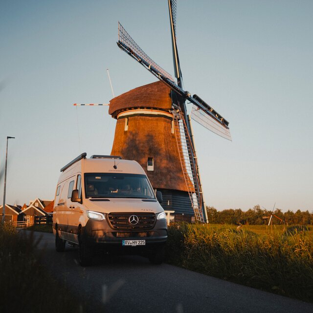 Grey camper van stands on a road, high grass grows on both sides of the road, behind the camper van there is a large windmill and further in the background you can see small houses