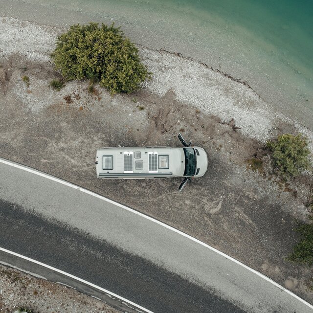 Grey camper van from above, standing on gravel next to a lake at the shore, next to the gravel there is a small road and on the gravel bed there are three green trees, the doors of the camper van are open