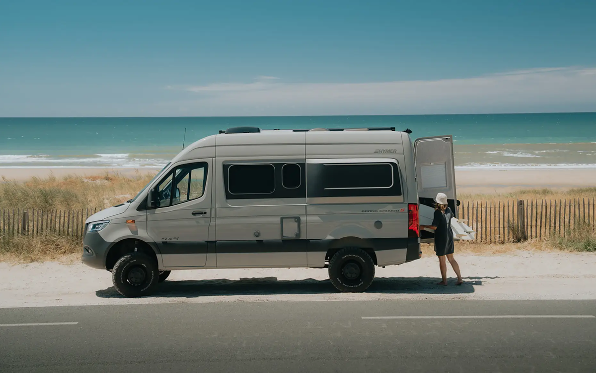 Side view of a stone grey campervan with black design accents and black all-terrain tyres, parked by the roadside directly at the beach with turquoise blue sea in the background under a bright blue sky. A person stands at the open rear door holding a surfboard.