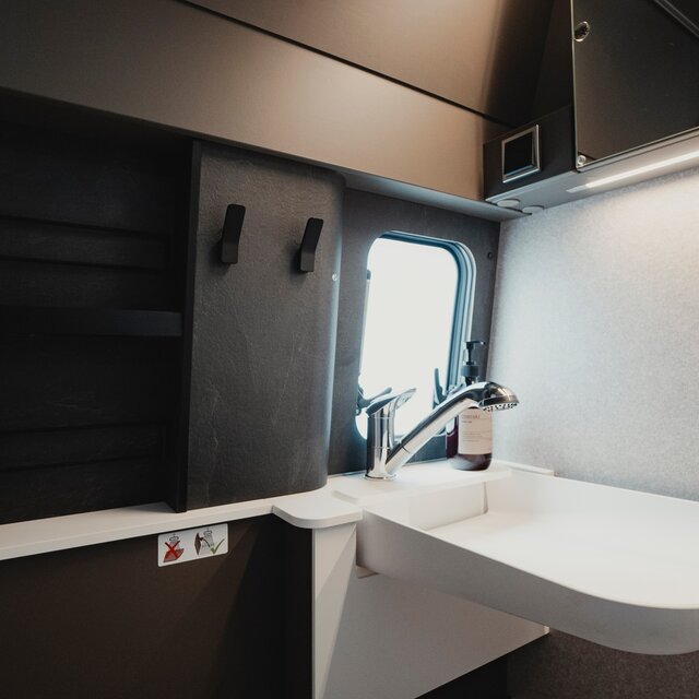 Sink in a bathroom of a camper van with silver faucet, next to it on the left you see a storage shelf in dark grey marble look and two black towel hooks