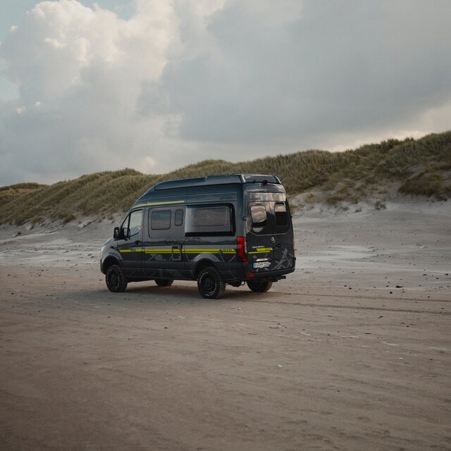 Black camper van with neon green special decals driving over sand, dunes visible in the background