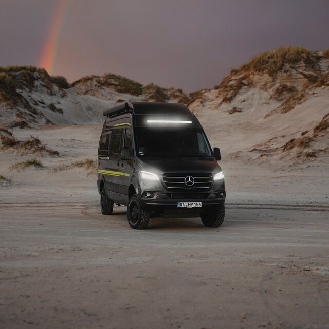 Black camper van with neon green special decals standing in the sand, dunes visible in the background, headlights and light bar shining in the twilight