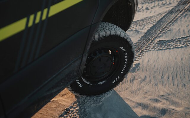 Black tire with white All-Terrain T/A lettering on a dark camper van standing on sand