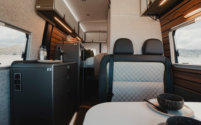 Living area in a modern camper van, set table and seating area with dark leather, kitchenette with cooking utensils, fridge and overhead storage cabinets, in the background you see a made bed