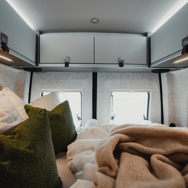Sleeping area in a camper van with made-up bed, green decorative pillows, and light overhead cabinets