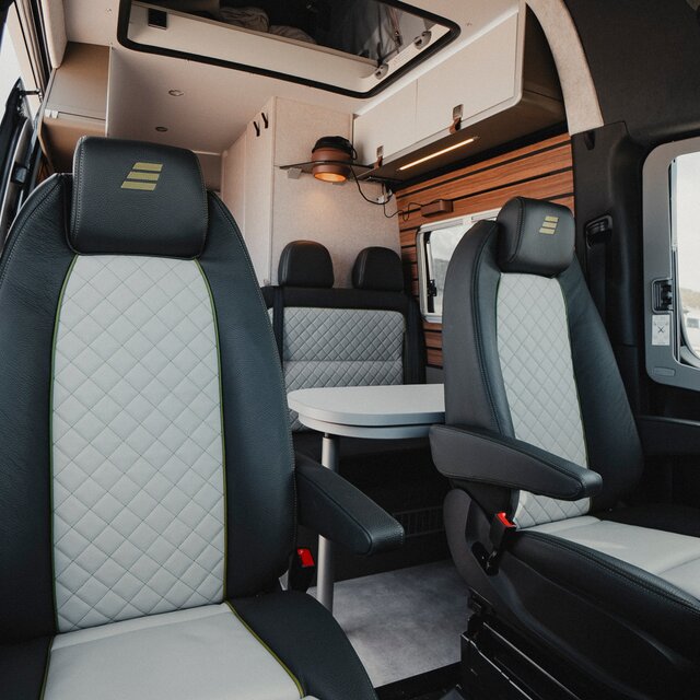 Two driver seats in a camper van with black-grey leather upholstery, in the background is the same colored seating group with table in front and above the light overhead cabinets as well as the brown pendant lamp