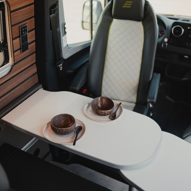 Set table in a modern camper van, the driver seats with grey-black leather are turned to the table, bottom left in the image you see the seating group and top left the side window