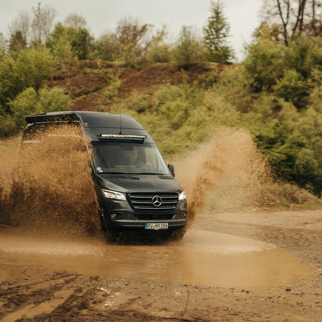 Camper van drives through a large puddle in an offroad park, splashing, with trees and dirt mounds in the background