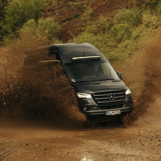 Camper van drives through an offroad park, it is dirty, with trees and dirt mounds in the background