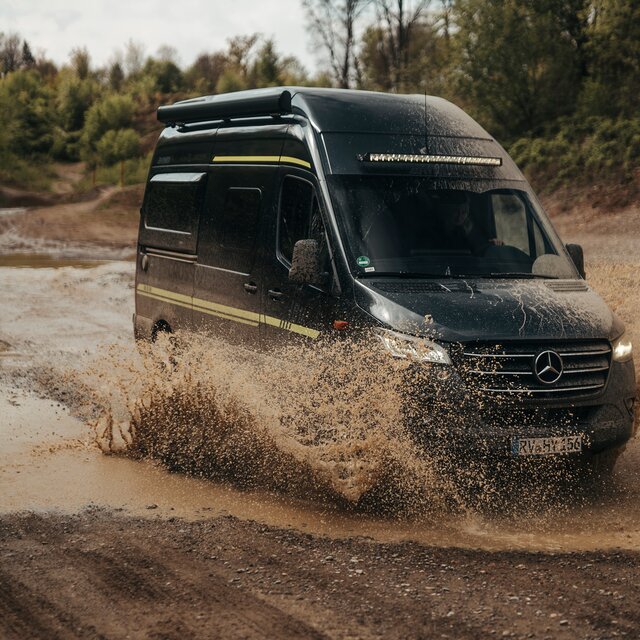 Camper van drives in the distance through an offroad park, with trees and dirt mounds in the background and a puddle to the left of the camper van