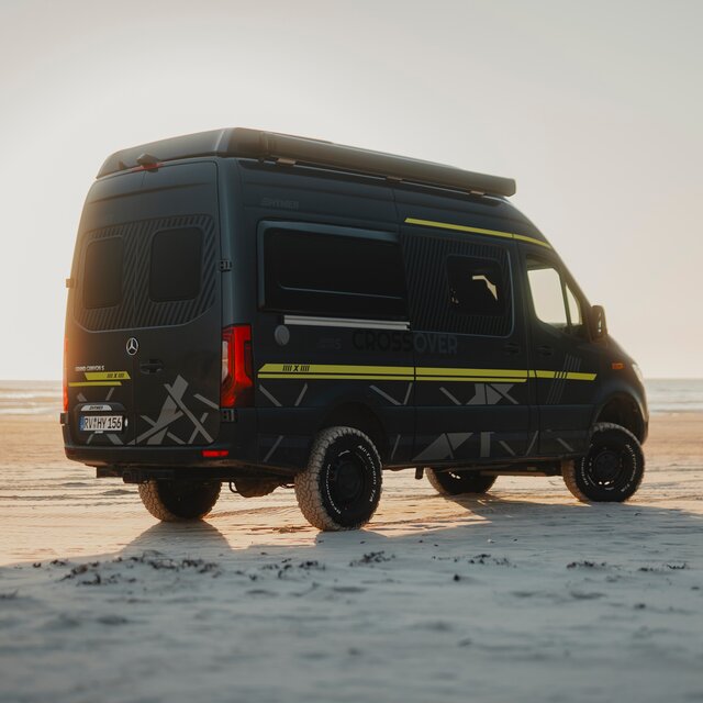 A part of the black grille of a black camper van, a grey 4x4 decal and the right headlight are visible, with dunes in the background