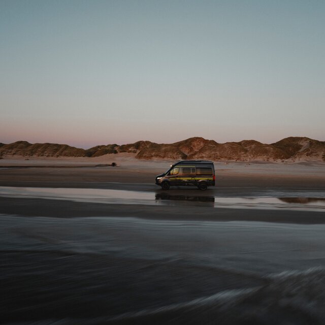 Black camper van with neon green decals drives sideways over sand, with dunes in the background