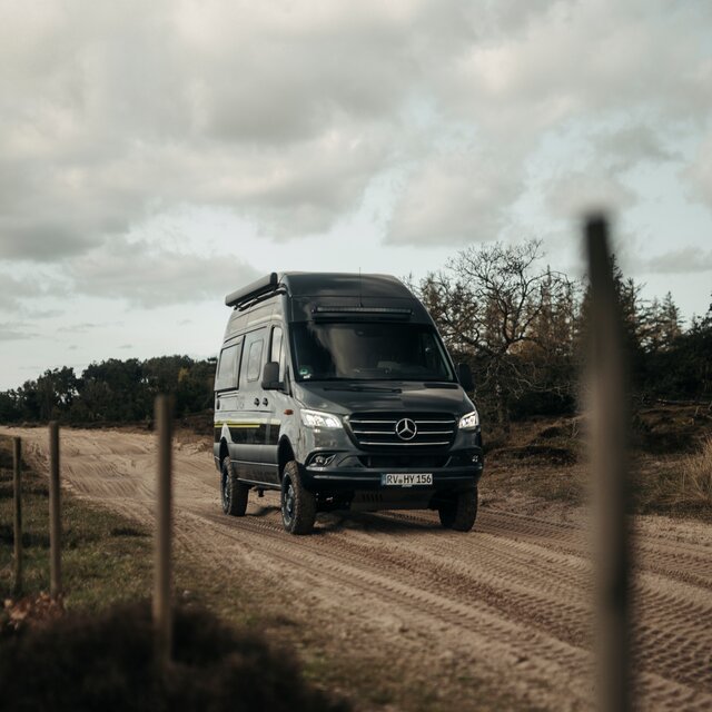 Black camper van drives over a muddy road, with trees in the background and a fence with wooden posts to the left of the road