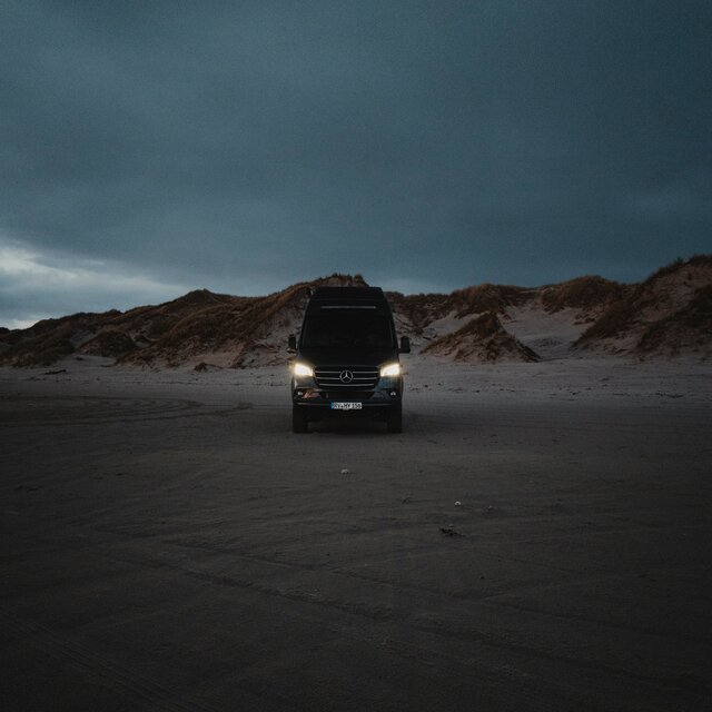 Black camper van from the front, standing on sand with dunes in the background, it is dark and the headlights are on