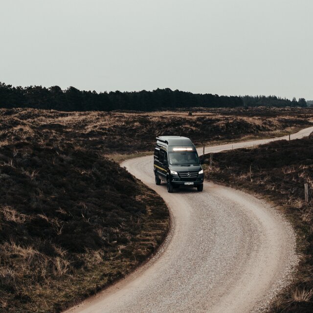 Black camper van drives over a rocky road, with higher grass on both sides of the road, a forest is visible in the background, the headlights and LED light bar above the windshield are on
