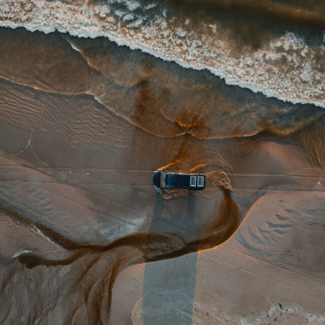 Black camper van drives over the beach, seen from above, driving parallel to the sea