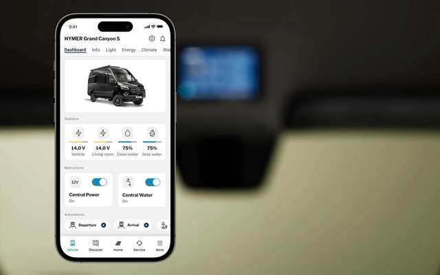 Display of a mobile phone screen showing the content of an app for controlling a campervan. The phone is depicted in front of a blurred campervan interior.
