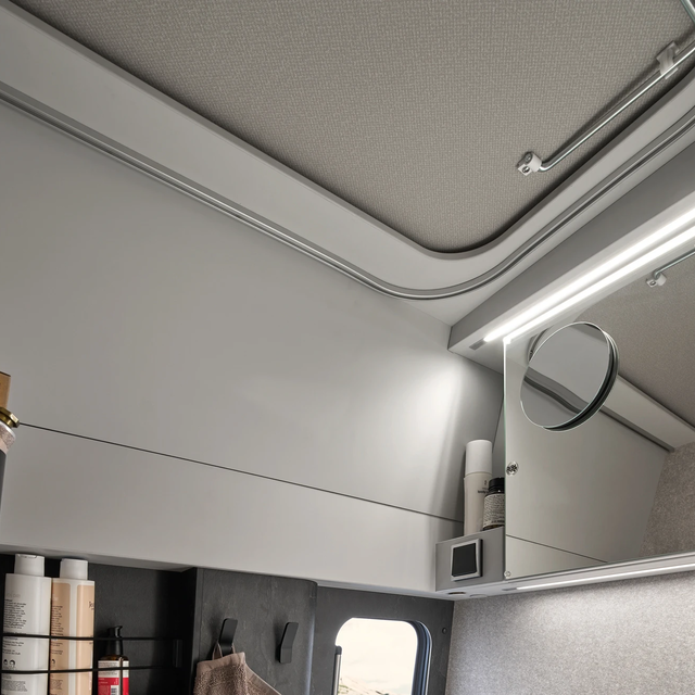 Close-up of a campervan bathroom, showing storage compartments with cosmetic products and on the right a large mirror with an attached round makeup mirror.