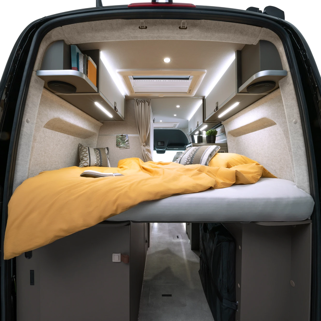 View through the open rear doors into a modern campervan. At the back of the vehicle is the bed with yellow bedding on it.