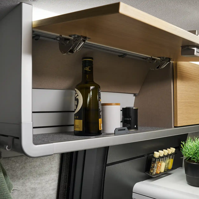The roof storage cabinet above the kitchen of the HYMER Grand Canyon S in the Pearl Grey style world is open. Oil and spices are visible inside.