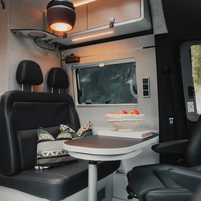 Living area in a modern camper van, set table with seating area with dark leather, the two driver's seats are turned towards the table, above the seating area are roof storage cupboards and a brown pendant lamp