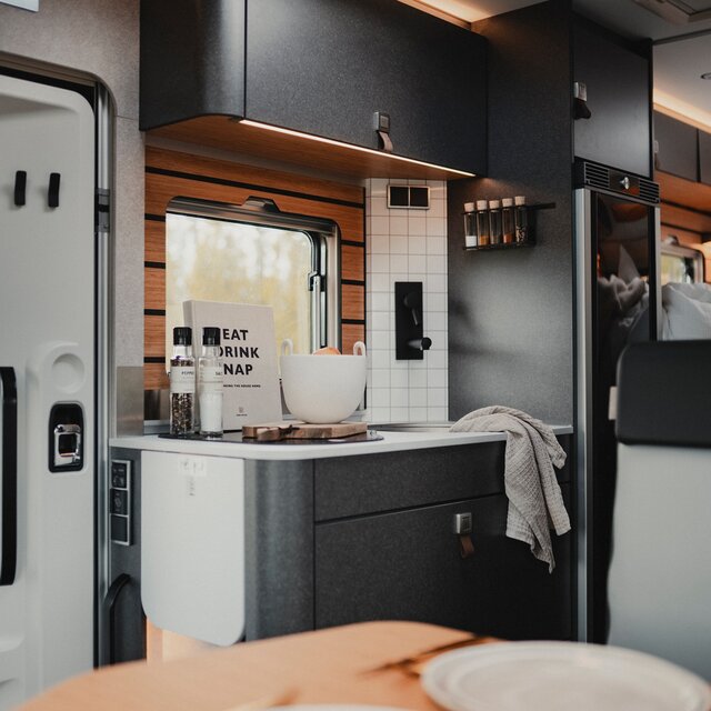 Interior of a motorhome, entrance door, kitchen unit with cooking utensils and countertop extension, foreground shows part of the set table and the back bench of the seating group