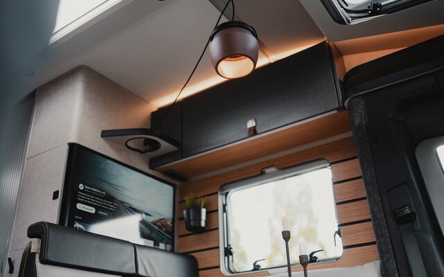 Dark overhead cabinet above the seating area in the living space of a motorhome also visible are the TV flat screen, side window, pendant light on the ceiling, headrest of the seating area
