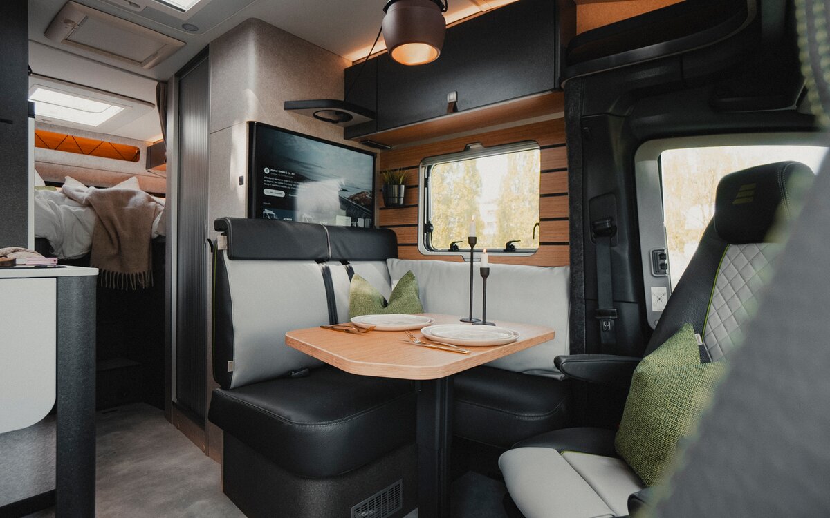 Living area of a motorhome with a view of a modern seating area with a dark leather-fabric combination, set table, TV flat screen, side window and overhead cabinet