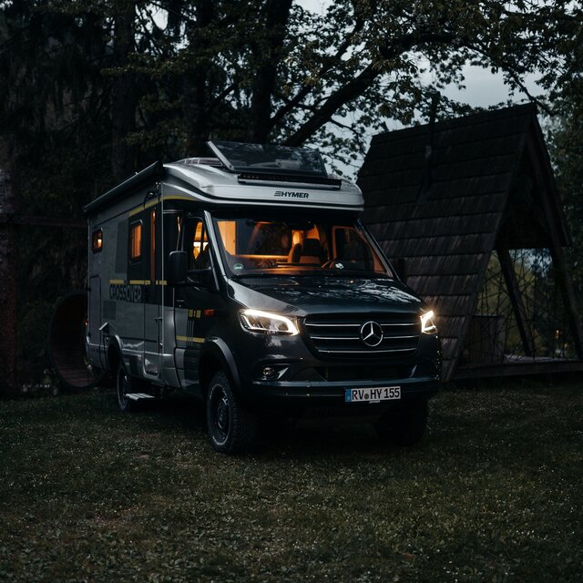 Semi-integrated motorhome stands in a small forest next to a pointed wooden hut in the dark and the light is on inside the motorhome