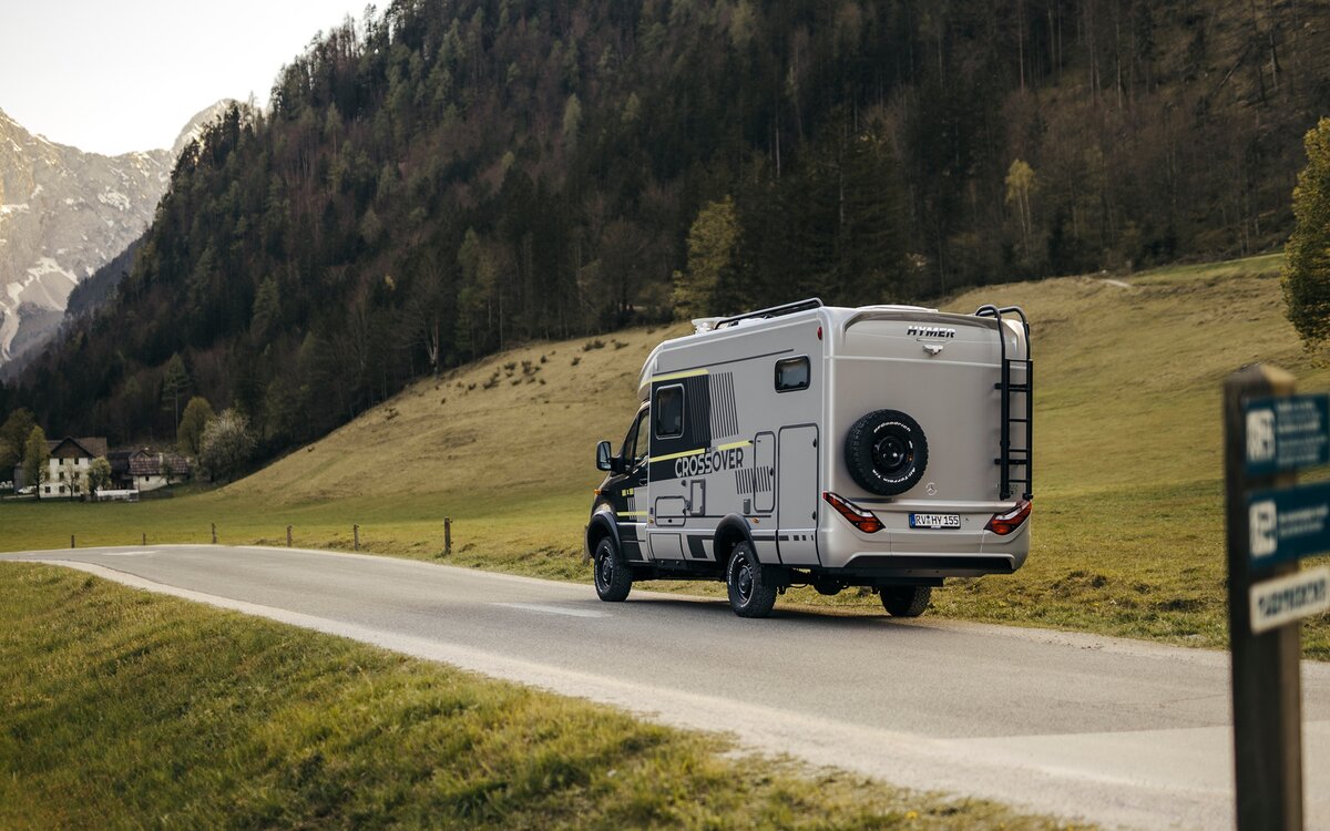 Semi-integrated motorhome drives on a country road with a mountain in the background, you can see the side and rear of the motorhome with a black ladder and a black spare wheel hanging from it