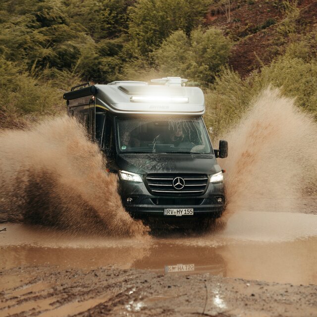 Semi-integrated 4x4 motorhome drives through a large puddle in an off-road park, puddle splashes, trees and dirt mounds are visible in the background