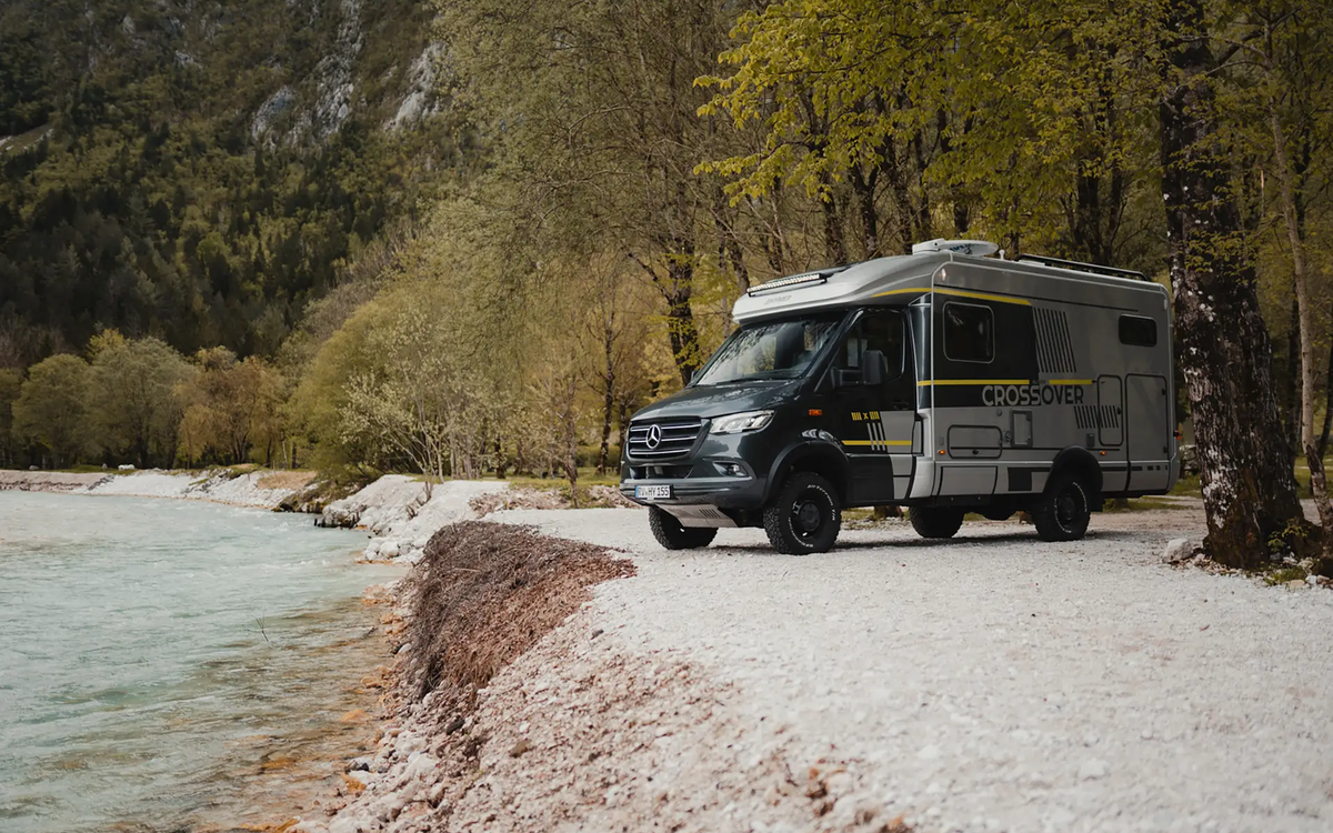 Semi-integrated 4x4 motorhome stands sideways on gravel by a riverbank with trees in the background