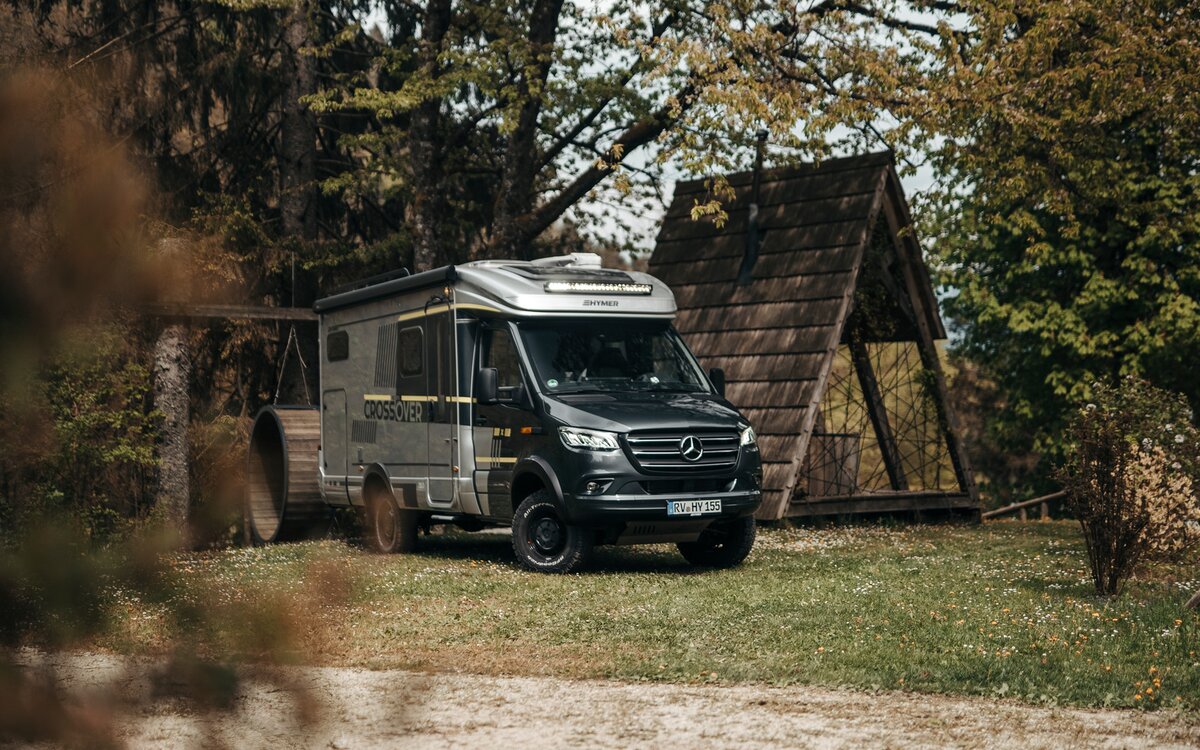 Semi-integrated motorhome seen from an angle in front, standing in a small forest next to a pointed wooden hut