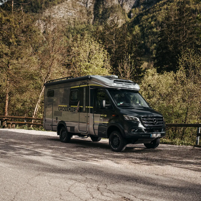 Semi-integrated motorhome seen from the front at an angle, standing by a roadside, in the background, you can see a wooden railing and a rock face with trees