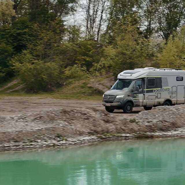 Semi-integrated motorhome, driving on gravel through an off-road park along a turquoise blue body of water, in the background, you can see tree-covered gravel hills
