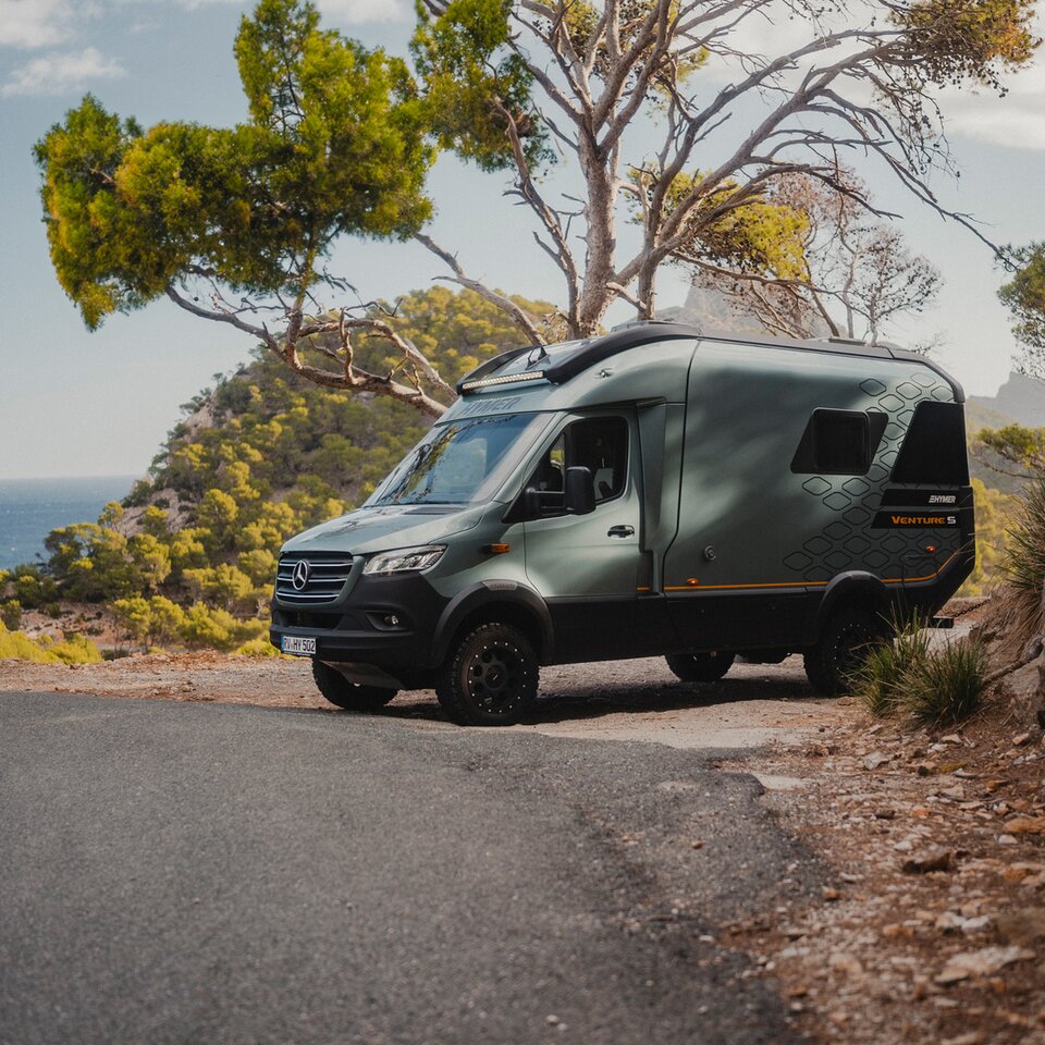 Utsikt från den främre sidan av Venture S, parkerad vid vägkanten, omgiven av berg, träd och havet.